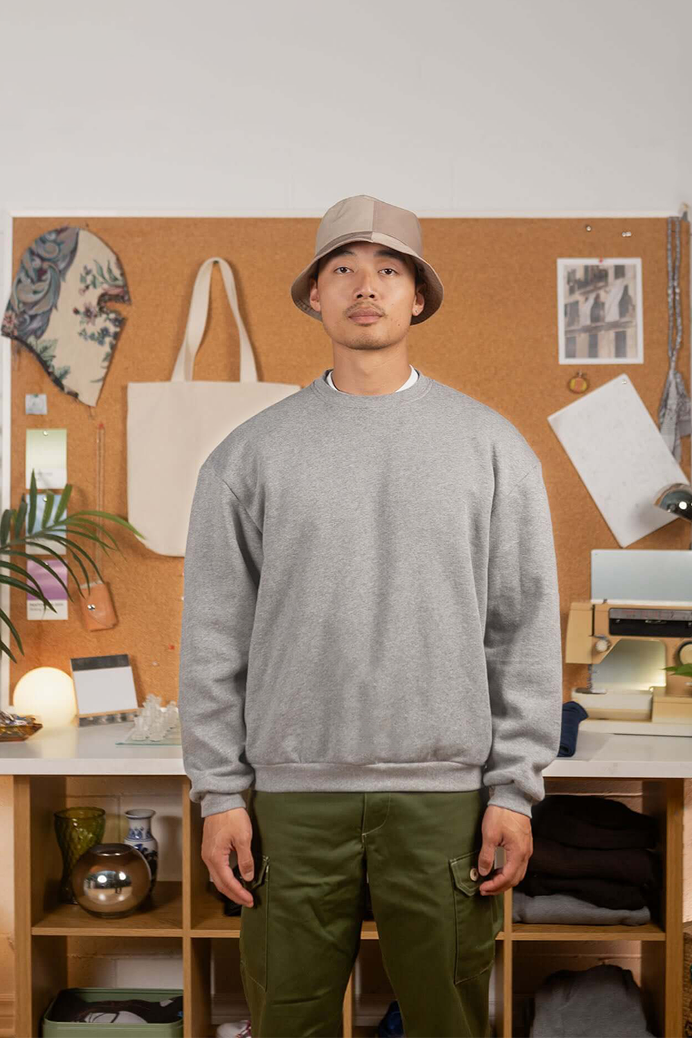 Man wearing a grey sweatshirt and green pants standing in front of a cork bulletin board with various items.