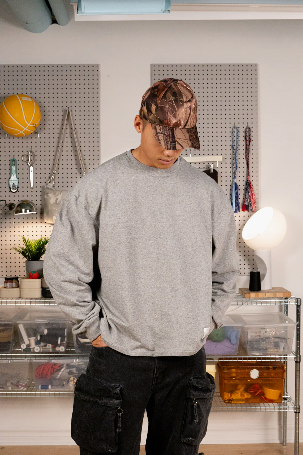 Person wearing a grey boxy long sleeve shirt and black pants in a room with shelves and decor.