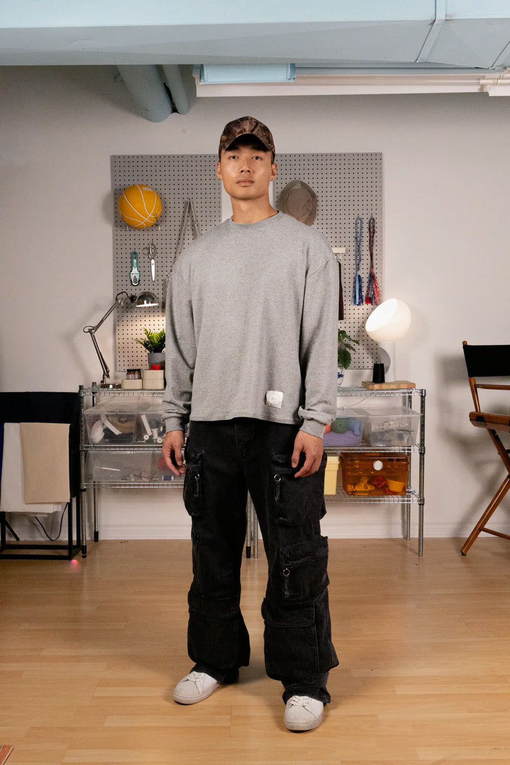 Person wearing a grey boxy long sleeve shirt and black pants standing in a room with a pegboard wall.