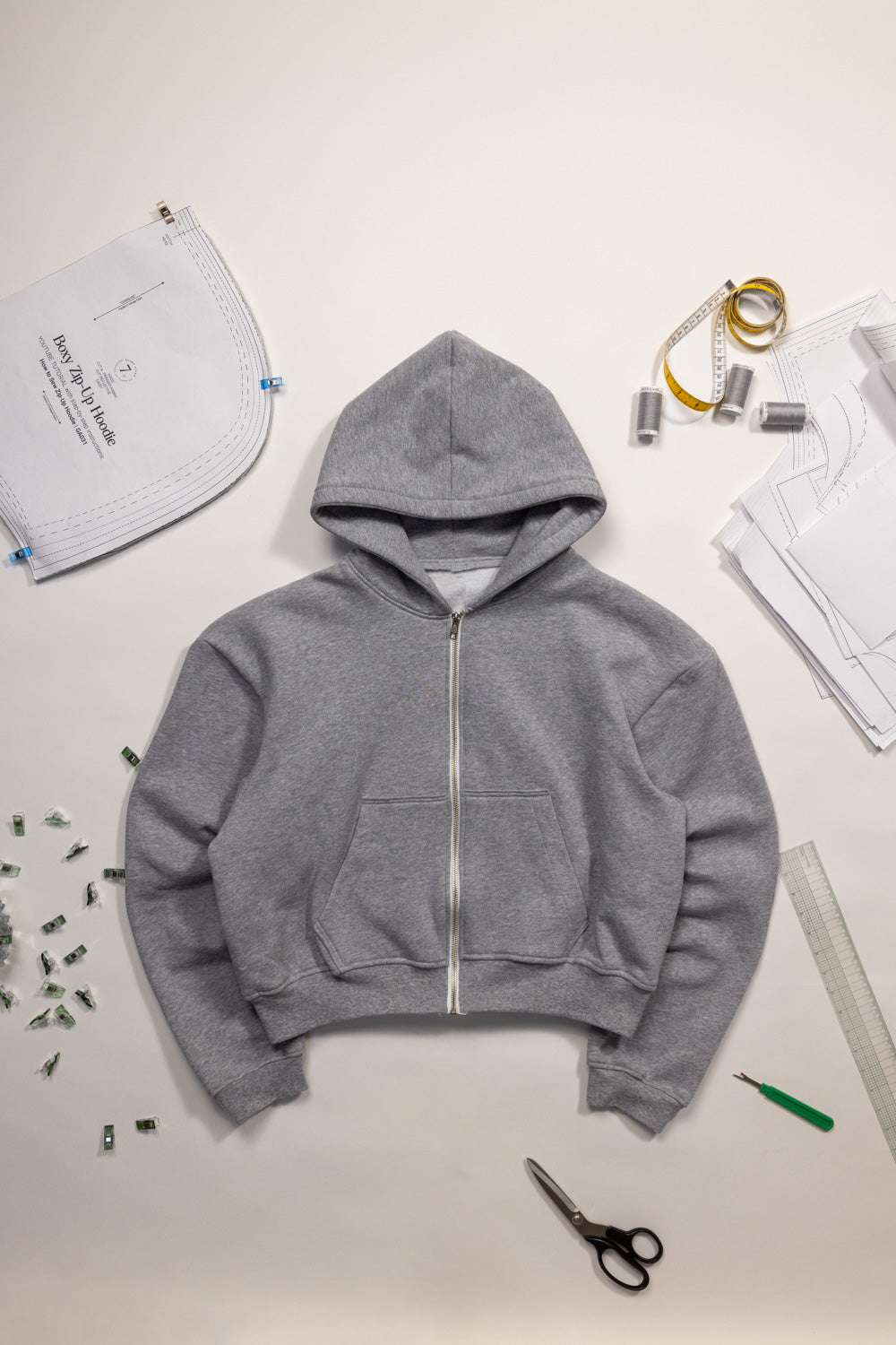 Gray boxy zip-up hoodie on a white surface with sewing tools and materials