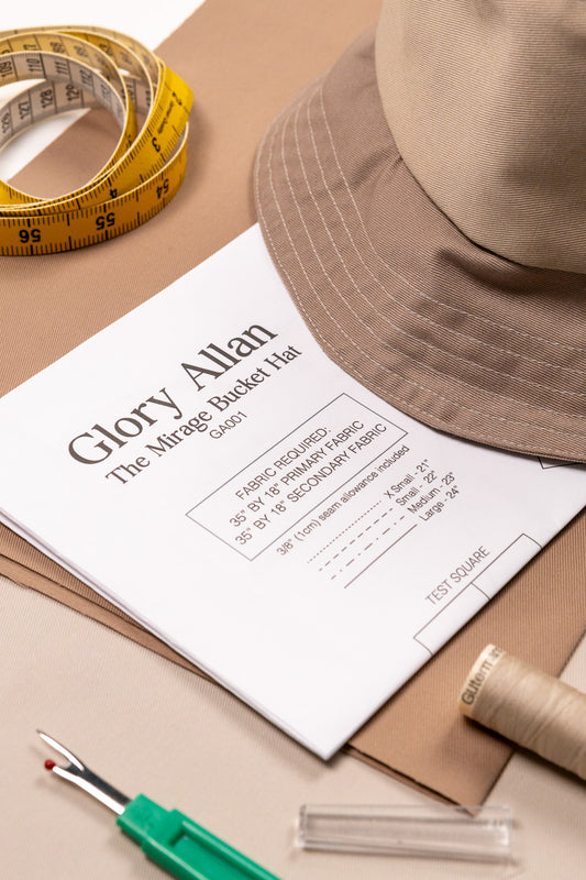 Brown bucket hat on a piece of paper with text, accompanied by a measuring tape, pen, and spool of thread on a beige background.