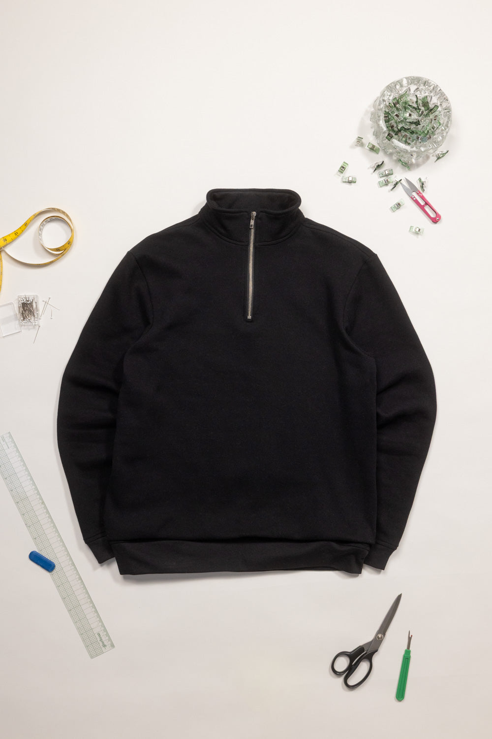 Black half-zip sweatshirt on a white surface with sewing tools.